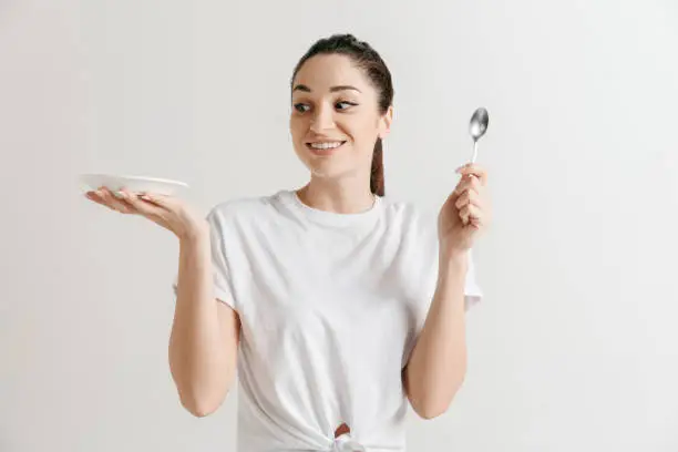 Young fun crazy brunette housewife with spoon isolated on white background. Housekeeper woman holding white empty plate. Copy space advertisement.