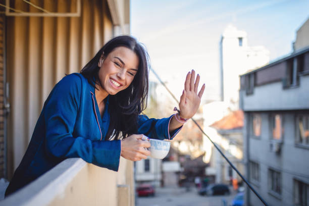 Smiling neighbor waving while enjoying a cup of coffee Cute and beautiful young woman enjoying a cup of coffee outdoors in the morning on a building terrace. neighbour stock pictures, royalty-free photos & images