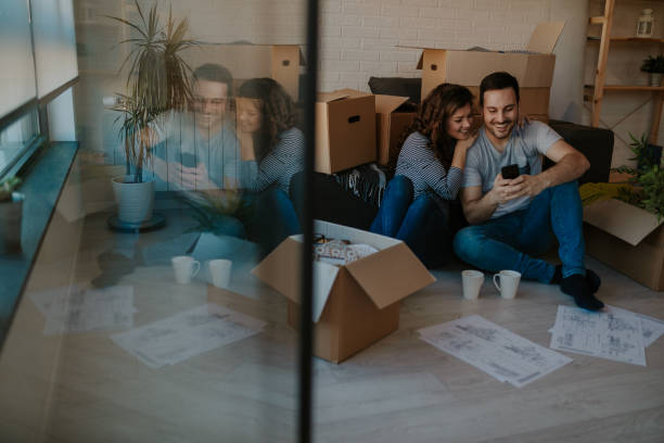 Young happy couple sitting, relaxing and looking at the mobile phone surrounded by unpacked boxes. stock photo