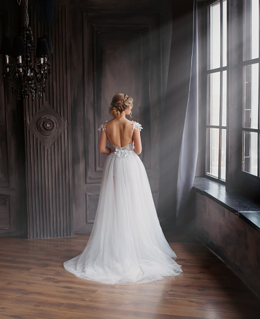 amazing lady in long white adorable expensive light dress with train and open back stands with back to camera, girl with blond hair posing in spacious room with large windows with sunlight. no face.