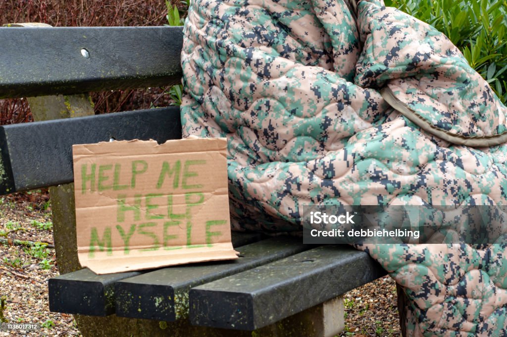 Homeless Person on Park Bench Homeless person wrapped in camouflage blanket sitting on park bench with handwritten cardboard sign asking for help Homelessness Stock Photo
