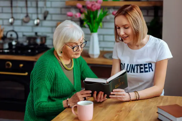 Having heart-to-heart talk. Beautiful grey-haired retired woman wearing green sweater in glasses having heart-to-heart talk with young-adult fair-haired understanding lady at kitchen.