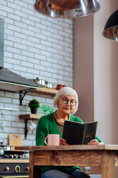 istock Elderly female busy with mindful reading of Bible at kitchen 1136018657
