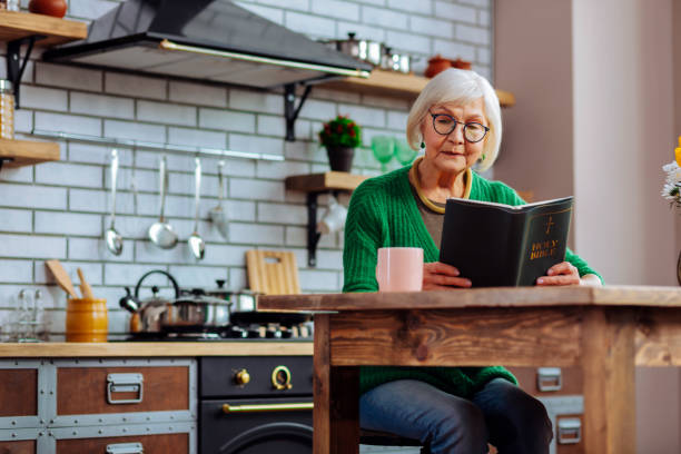 istock Old madam enjoying reading of Bible book at kitchen table 1136018658