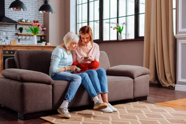 Daughter and mother spending time together. Elderly charming grey-haired female in light-blue sweatshirt and jeans sitting on sofa with her young-adult bewitching blonde daughter with phone in hands.