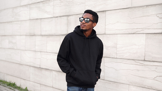 Portrait african man in black hoodie, sunglasses walking on city street looking away over gray brick wall background