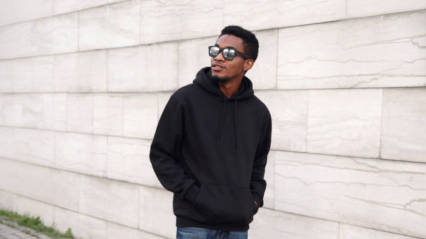 Portrait african man in black hoodie, sunglasses walking on city street looking away over gray brick wall background stock photo
