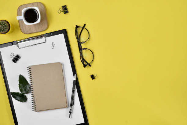 istock Office desk workspace with blank clip board, office supplies, pen, cactus, green leaf, coffee cup on a wooden stand and eye glasses on yellow background. Flat lay, top view blog concept 1136023154