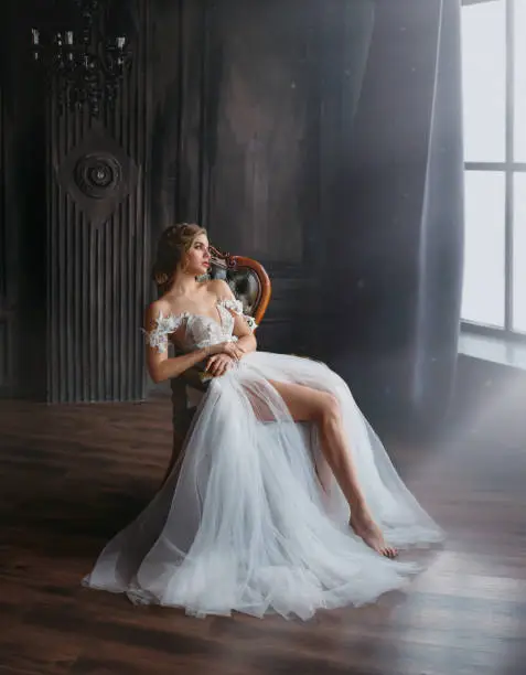 Photo of majestic and proud princess girl in white chic oriental white silver dress tired sitting on chair, lady shows off her slender leg and waiting for prince, gentle stylish image of graduate 2019