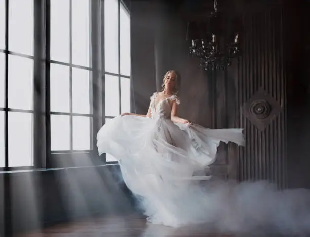 Photo of sweet and gentle girl with fair skin and blond hair dances alone in silence in an old castle, spirit of abandoned medieval building whirls in white smoke and magical thick fog in rays of sun