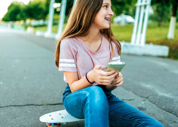 12-16 세의 어린 소녀 여 학생, 거리에서 그녀는 공원에서 여름에 스케이트 보드에 앉아. 전화의 손에, 온라인 응용 프로그램, 인터넷 소셜 네트워크, 웃는 기 뻐 웃음 행복. - 12 16 years old 뉴스 사진 이미지
