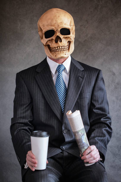 dead zombie hombre en azul marino rayas traje de negocios con corbata azul claro contra un fondo gris usando una máscara de cráneo humano aterrador sosteniendo un periódico y taza blanca de café-imagen - enzo fotografías e imágenes de stock