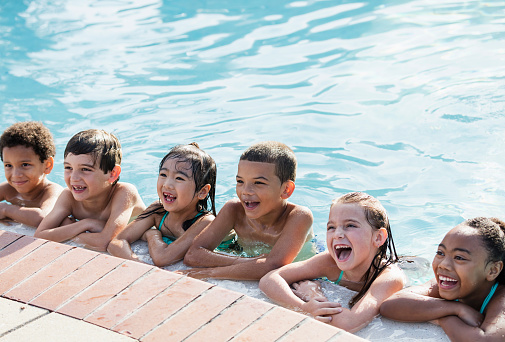 A multi-ethnic group of six children, 6 to 9 years old, having fun at a swimming pool, in the water hanging onto the side, in a row, watching something, shouting and laughing.