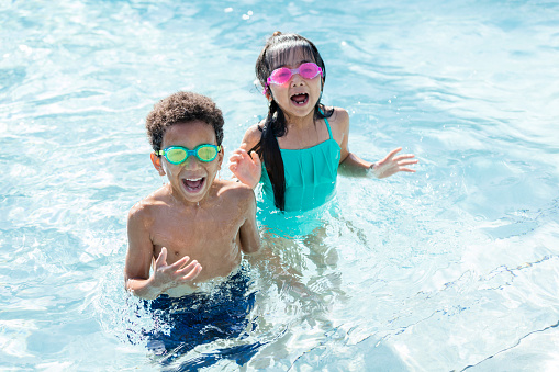 Two multi-ethnic children, 6 years old, having fun in a swimming pool. They are standing waist deep in water, looking up at the camera, shouting and laughing. The boy is mixed race African-American and Caucasian and the girl is Japanese and Filipino.