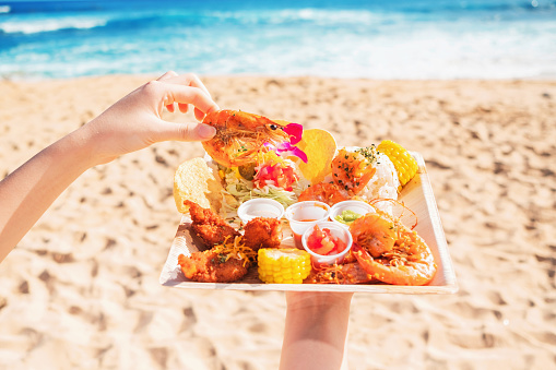 Holding food plate of shrimp, rice, chicken, corn and sauces on the beach