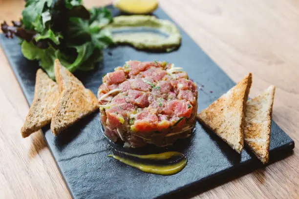 Close up Spicy Bluefin Tuna Tartare with sour and spicy sauce. Served with toast and salad on black stone plate.