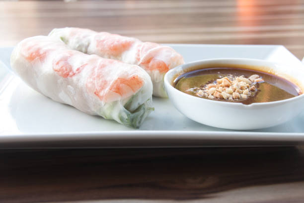 A close-up of fresh Thai spring rolls filled with crisp lettuce, rice noodles, and vibrant vegetables, delicately wrapped in rice paper. The rolls are served with a tangy dipping sauce in a glass bowl, presented on a traditional blue-patterned plate. This healthy and flavorful appetizer embodies the light, refreshing, and authentic essence of Thai cuisine, making it a visually appealing and satisfying dining experience.