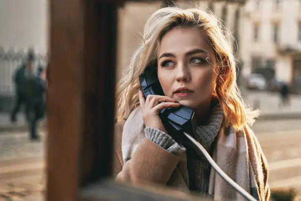Photo of Young beautiful blond woman making an important call in a vintage public phone booth on a sunny evening
