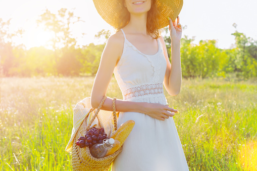 Portrait of beautiful young woman holding basket with bottle of water, baguette and grape for picnic. Romantic girl in white dress and fashionable hat is smiling on field at sunset. Provence style.