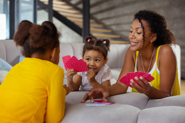 Happy brunette girl looking at her sister Playful mood. Young dark-skinned female keeping smile on her face while taking free card family playing card game stock pictures, royalty-free photos & images