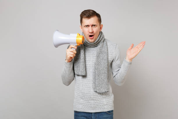 istock Perplexed young man in gray sweater, scarf hold megaphone spreading, pointing hand aside isolated on grey background. Healthy lifestyle people sincere emotions cold season concept. Mock up copy space. 1136177530