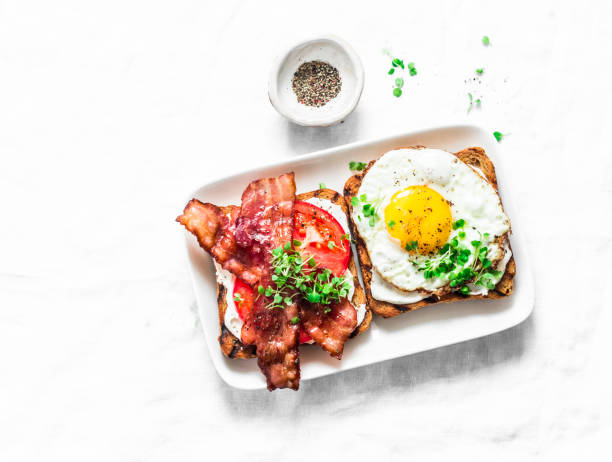 A plate of English breakfast, scrambled eggs, fried bacon and wheat toast on a dark wooden table, cutlery nearby. Close-up.