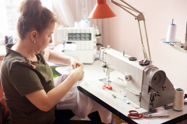 istock Woman stitching white dress with hands and needle. Sewing machine, scissors, pins, threads, table lamp on background. 1136190208