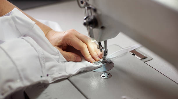 istock Female hands at sewing process and repairing white fabric on professional manufacturing machine. Close up top side view. 1136190214