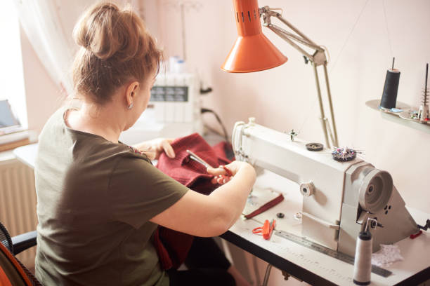 istock Tailor woman sitting on workplace in atelier and using scissors for cutting off cloth piece in sewing process. Back view 1136190232