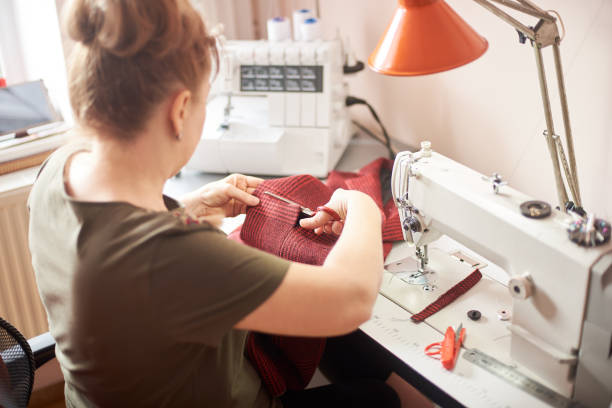 istock Tailor woman sitting on workplace in atelier and using scissors for cutting off cloth piece in sewing process. Back view 1136190236