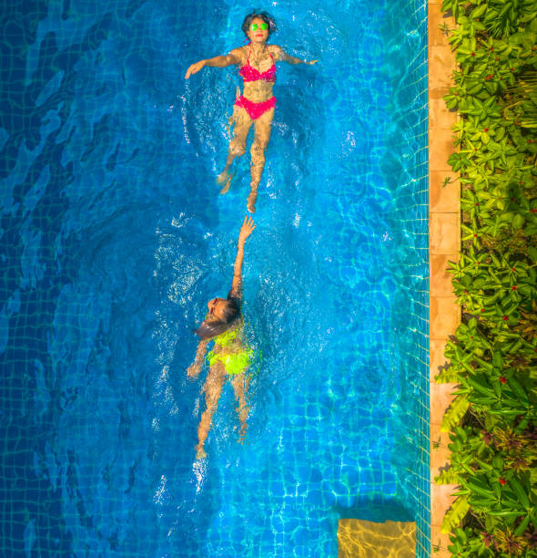 aerial top view above swimming pool. woman in yellow bikini and woman in red bikini are swimming \