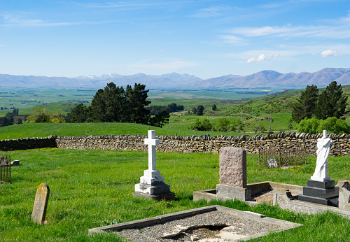 View across old cemetery with broken headstones to expansive rural fields and distant hills in Central Otago New Zealand