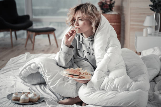 Sad and lonely woman eating burger and French fries in the bed Burger and fries. Sad and lonely blonde-haired young woman eating burger and French fries in the bed relationship breakup stock pictures, royalty-free photos & images