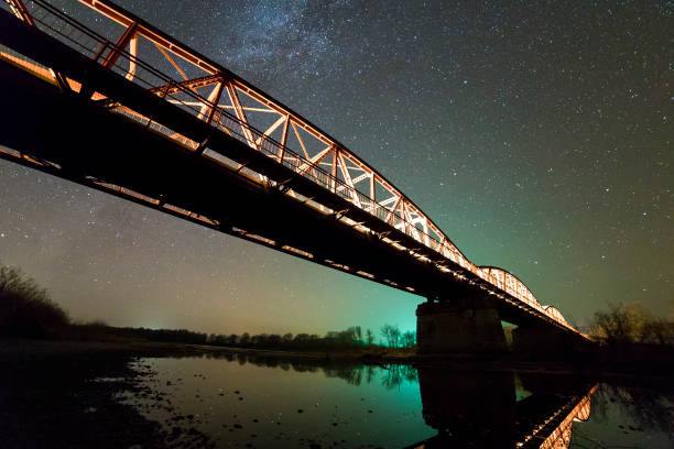 istock Illuminated metal bridge on concrete supports reflected in water on dark starry sky with Milky Way constellation background. Night photography concept. 1136226410