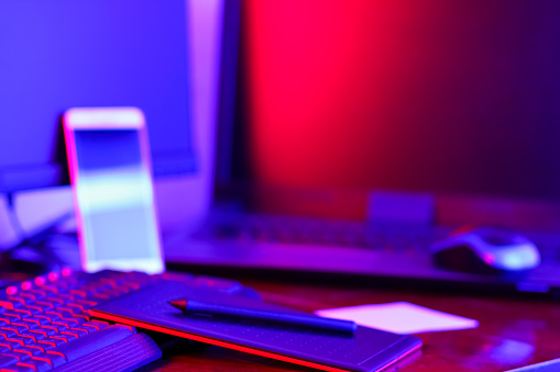 Workplace digital designer. Freelancer workplace in neon light. Computer, graphics tablet and smartphone on the table. Place for creativity in blue pink light