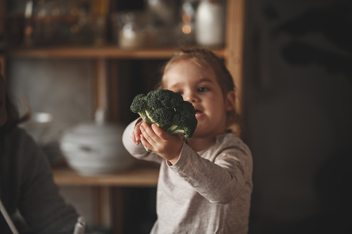 Two year old cute baby girl is sitting on the kitchen counter, holding fresh broccoli.
