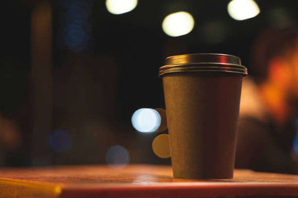 A black paper take away coffee cup on a wood table at night with blurry background