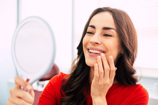 perfekt geehrt. fabelhafte frau in rotem pullover gekleidet, mit lockig langen haaren und eleganten händen hält einen spiegel für die suche durch die letzte arbeit der zahnarzt. - teenager dentist patient cheerful stock-fotos und bilder