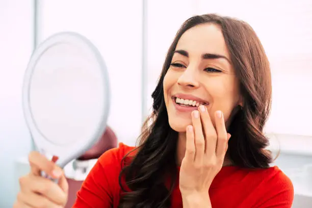 Photo of Honed to perfection. Fabulous woman clothed in red sweater, with curly long hair and elegant hands is holding a mirror for looking through the final work of dental doctor.