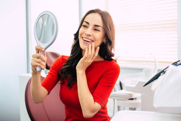 Long-term result. Nice looking girl in scarlet sweater is holding a mirror in her elegant hand and looking through the perfect  result of the dental work. stock photo