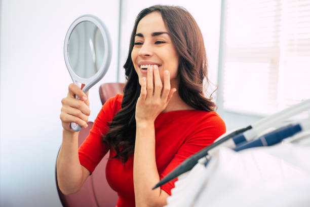 ultimatives ergebnis. atemberaubendes lächeln der freude schöne mädchen mit langen dunkelbraunen haaren, volle lippen, haselaugen und elegante hände mit einem spiegel in einem von ihnen vor hellem zahnzimmer. - teenager dentist patient cheerful stock-fotos und bilder