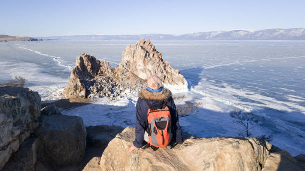 Girl traveler sitting on Cape Burhan and admires the rock Shaman on the island of Olkhon in Russia. Backpack over shoulders. Baikal in ice. A woman makes a trip to winter Siberia stock photo