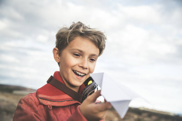 istock Smiling Boy Holding Paper Airplane While Standing Against Cloudy Sky 1136331659