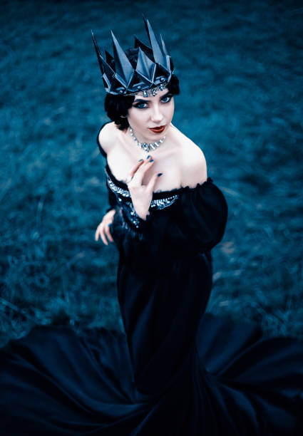 signora cupa con pelle bianca pallida e capelli scuri accanto all'erba congelata, la strega gira con corvo nero con lungo abito lussuoso e spalle aperte, immagine e trucco gotico, corona di metallo freddo e gioielli - women gothic style sensuality mystery foto e immagini stock