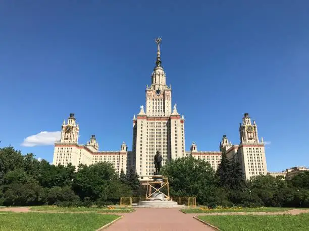 Moscow State University building and Lomonosov monument surrounded by green trees against the backdrop of a bright blue summer sky, Moscow, Russia