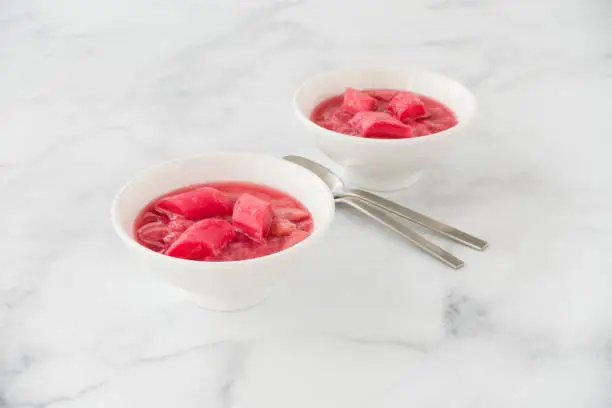 Homemade rhubarb compote, dessert, made from pink forced rhubarb, in two white porcelain bowls on white marble background.
