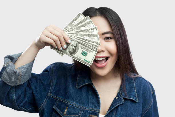 portrait of beautiful happy young asian girl in blue denim jacket standing, holding many cash dollars and covering half of face with toothy smile. - half smile imagens e fotografias de stock