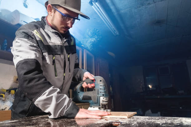 istock Young man with a beard in gray overalls and goggles by profession a carpenter works as an electric jigsaw in his home workshop. Cutting wooden parts 1136344187