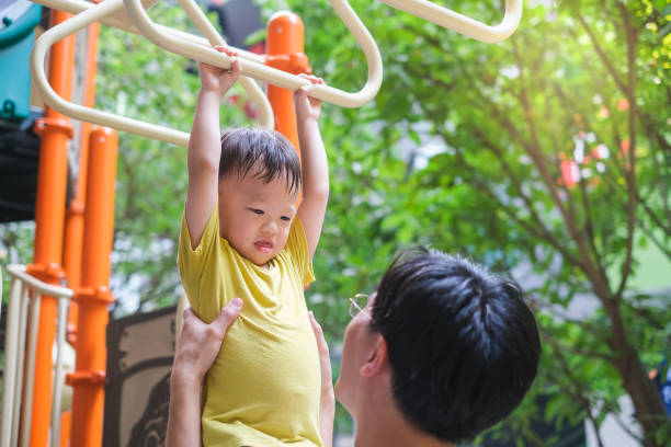 father and cute little asian 2 - 3 years old toddler baby boy child having fun exercising outdoor and dad help catch up on monkey bars equipmen - 2 3 years fotos imagens e fotografias de stock