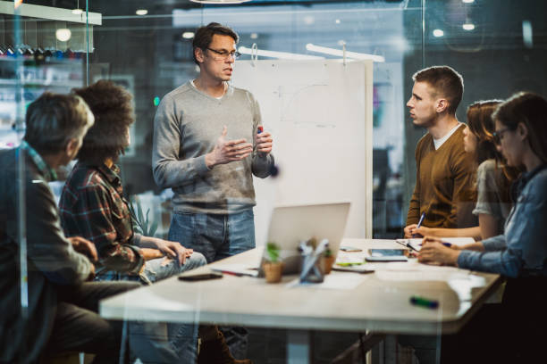 Mid adult businessman talking to his colleagues on presentation in the office. Mid adult manager talking while having presentation with his team in the office. The view is through glass. staff meeting stock pictures, royalty-free photos & images